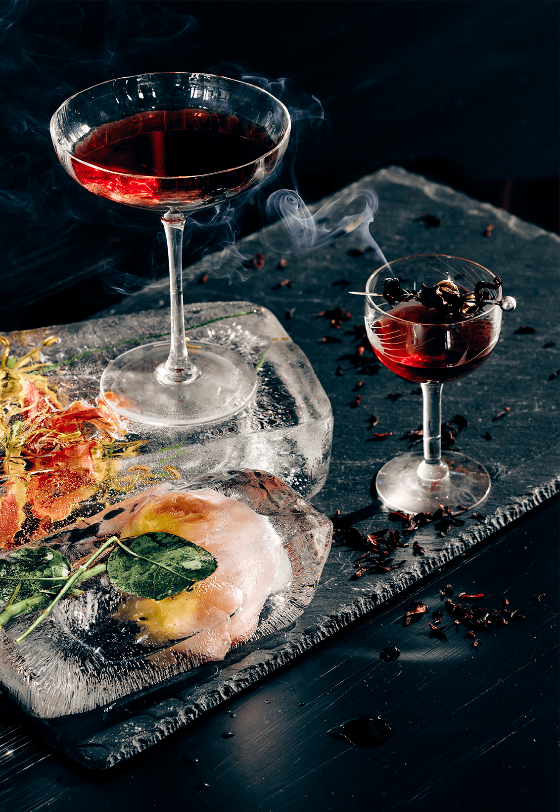 Two smoking crimson cocktails on botanicals and ice, dark and dramatic